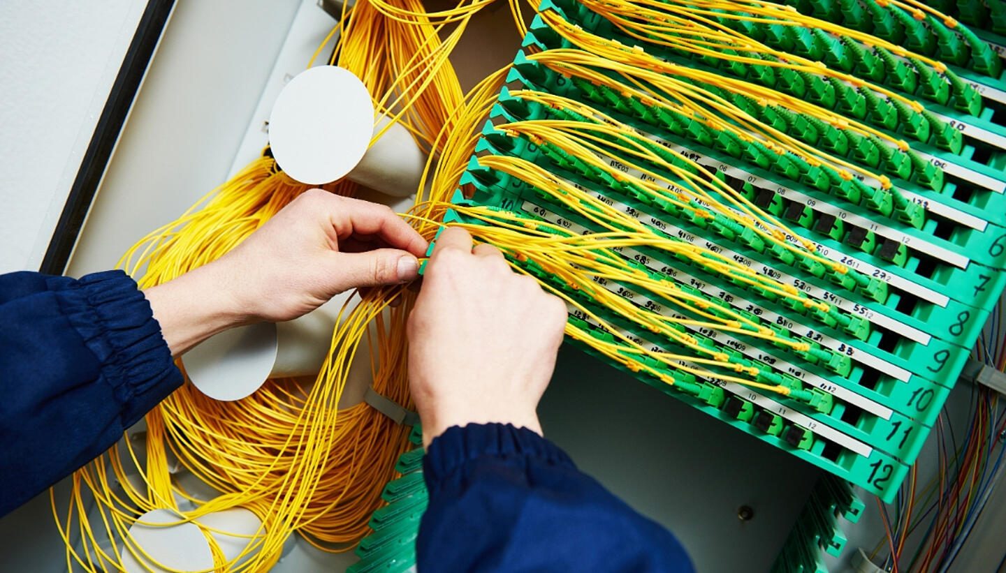 Zwei Hände adjustieren farbenfrohe Kabel in einer Schaltzentrale, damit der NE4 Ausbau gelingt.