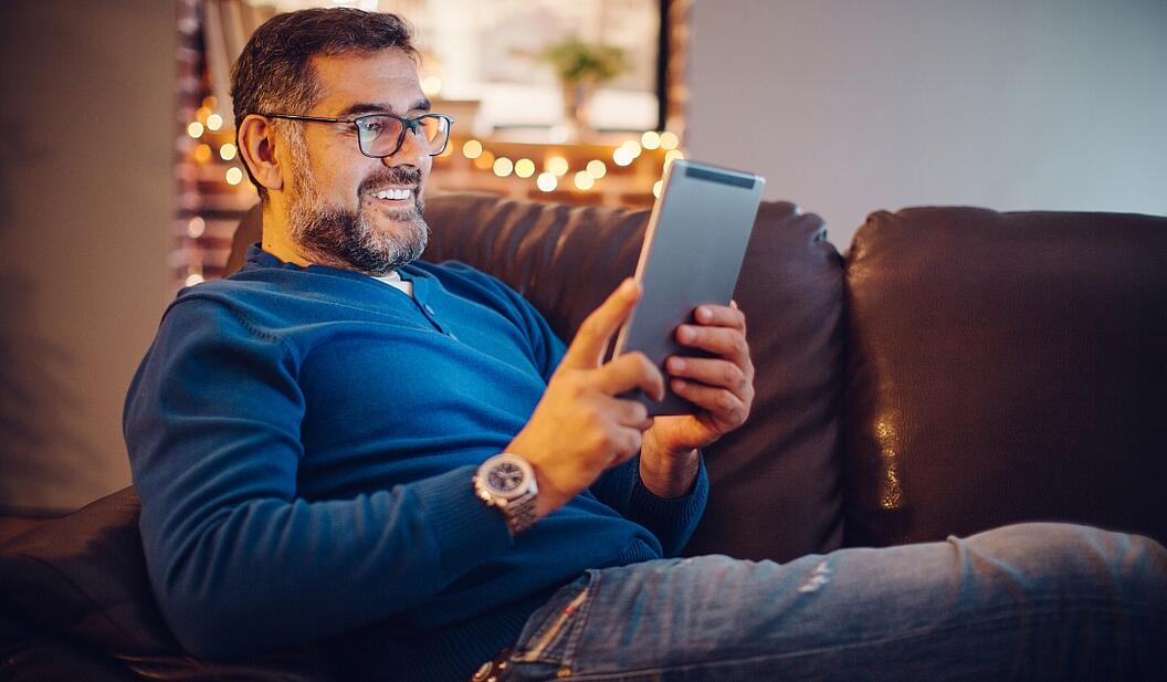 Ein Mann sitzt gemütlich auf einem Sofa und lächelt dank geglücktem NE4 Ausbau über etwas auf seinem Tablet, im Hintergrund eine Lichterkette.